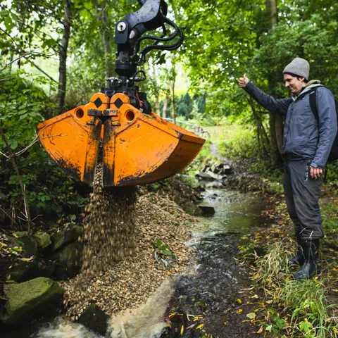 Eine große orangefarbene Baggerschaufel schüttet Kies in einen kleinen Bach, um das Flussbett zu verbessern. Ein Mann in wetterfester Kleidung, mit Gummistiefeln und Mütze, gibt mit einem Daumen-hoch-Zeichen ein Signal. Im Hintergrund stehen mehrere Personen in einer grünen, bewaldeten Umgebung und beobachten die Maßnahme.