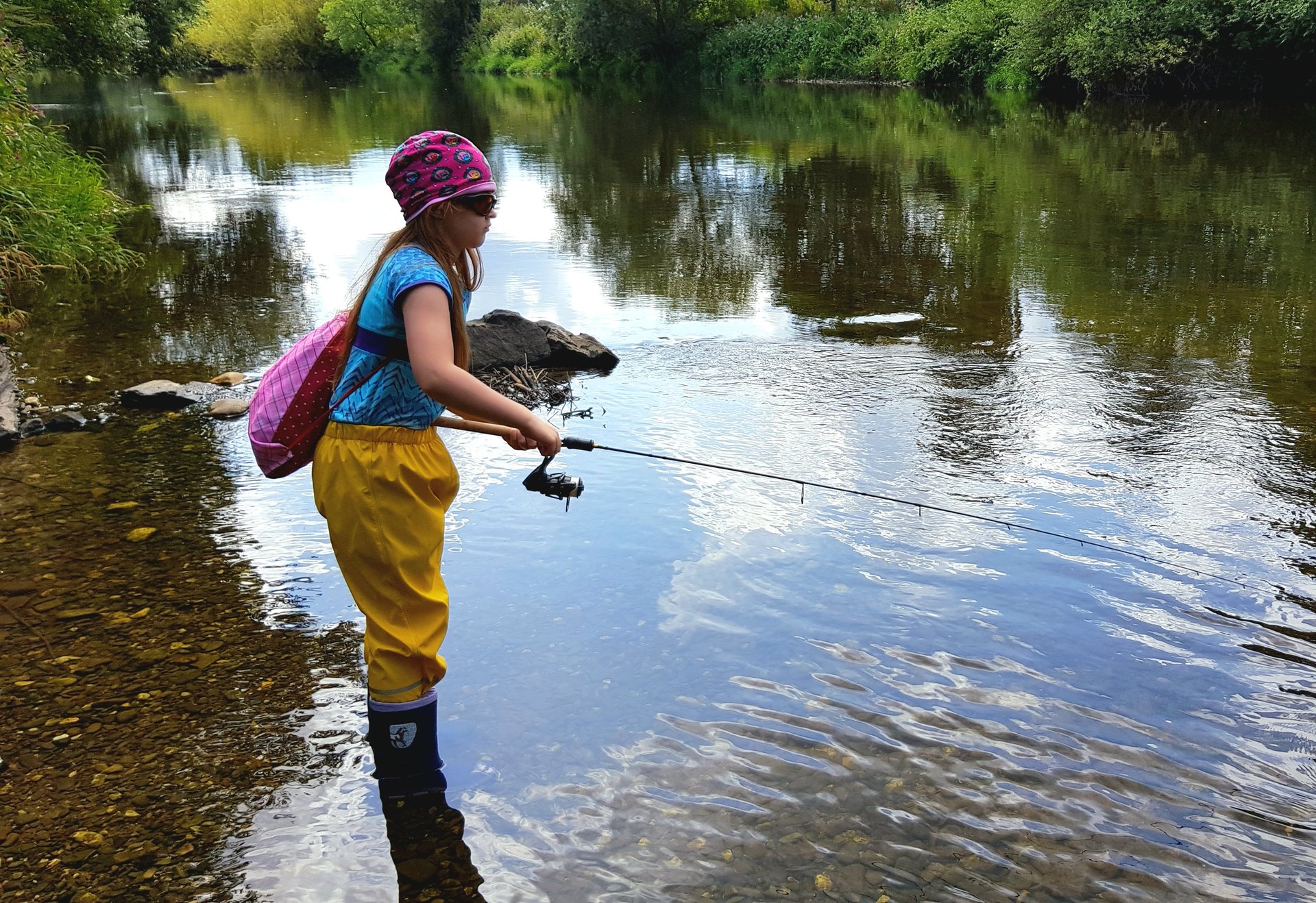 Ein Kind steht in einem flachen, klaren Fluss und angelt mit einer kleinen Angelrute. Es trägt eine bunte Mütze, eine Sonnenbrille, eine blaue Kurzarmbluse, gelbe wasserdichte Hosen und Gummistiefel. Auf dem Rücken trägt es einen pinken Rucksack. Die Umgebung ist von dichter grüner Vegetation gesäumt, und die Wasseroberfläche reflektiert den Himmel und die Bäume.