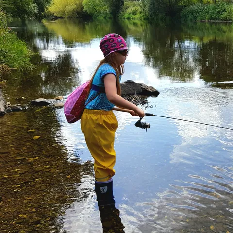Ein Kind steht in einem flachen, klaren Fluss und angelt mit einer kleinen Angelrute. Es trägt eine bunte Mütze, eine Sonnenbrille, eine blaue Kurzarmbluse, gelbe wasserdichte Hosen und Gummistiefel. Auf dem Rücken trägt es einen pinken Rucksack. Die Umgebung ist von dichter grüner Vegetation gesäumt, und die Wasseroberfläche reflektiert den Himmel und die Bäume.